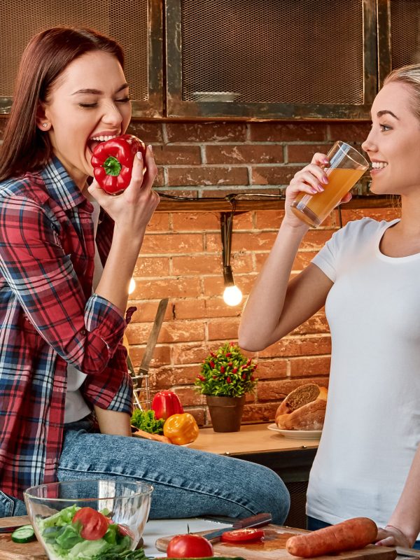 Portrait of two attractive women, having fun, while preparing salad. They are fully involved in the process. Dark-haired girl in checkered shirt sitting on the tabletop, she is going to bite a pepper. Her blonde friend in white T-shirt drinks fresh juice and looks at her