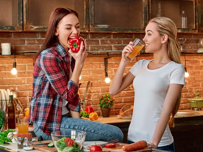 Portrait of two attractive women, having fun, while preparing salad. They are fully involved in the process. Dark-haired girl in checkered shirt sitting on the tabletop, she is going to bite a pepper. Her blonde friend in white T-shirt drinks fresh juice and looks at her